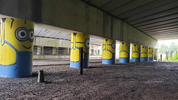 A row of eight painted stanchions under a motorway bridge, looking like yellow minions wearing blue dungarees and holding up the bridge