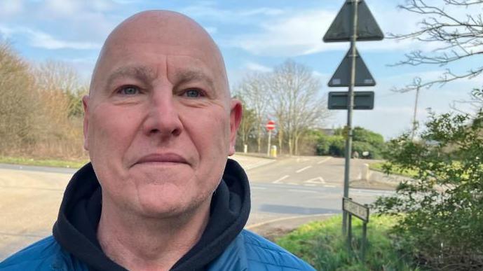 Paul Thomas, wearing a blue coat, and a black hooded top, looking straight at the camera, with a road sign behind him, and bushes and trees. 