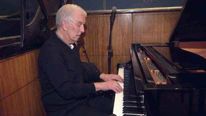 Bill Finlayson sitting at a piano. His hands are on the white keys. He is wearing a black jumper. He has white hair. A microphone is in the background in front of wood panelling.