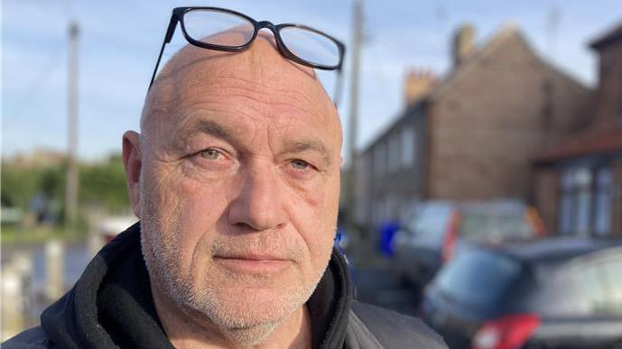 Johnny Whitfield smiles at the camera with black glasses that rest on his head. He is standing on a residential street which is blurred behind him.