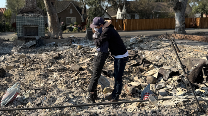 The couple hugging in the middle of the debris from the wildfires in LA. There is a house still standing in the background. 