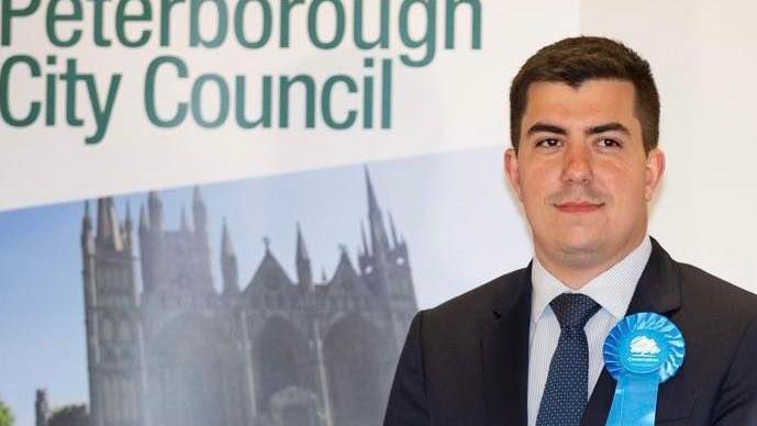 Rylan Ray stares at the camera. He wears a dark suit and a navy tie and has a Conservative party rosette. In the background is a Peterborough City Council sign with an image of the city's cathedral.