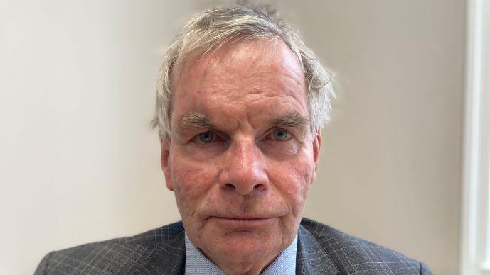 A close-up portrait of a man looking directly into the camera with a serious expression. He has short grey hair and is wearing a grey blazer and light blue shirt. The wall in the background is white.
