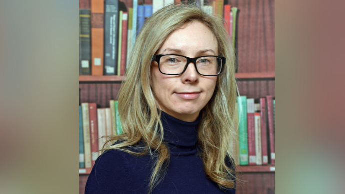 A headshot of Dr Yvonne McElarney with blurred bars at either side. She has long blonde hair and is wearing a pair of black rimmed glasses. She is wearing a navy turtleneck jumper.
