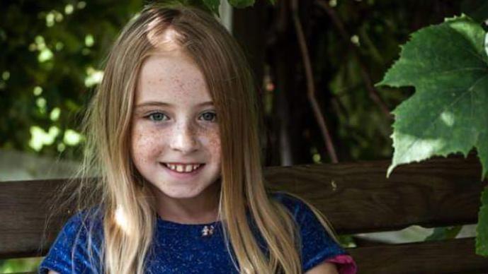 Cimarron Thomas is staring into the camera. She has dark blonde hair and blue eyes. She is wearing a blue dress with short sleeves. She is sitting on a bench with trees in the background