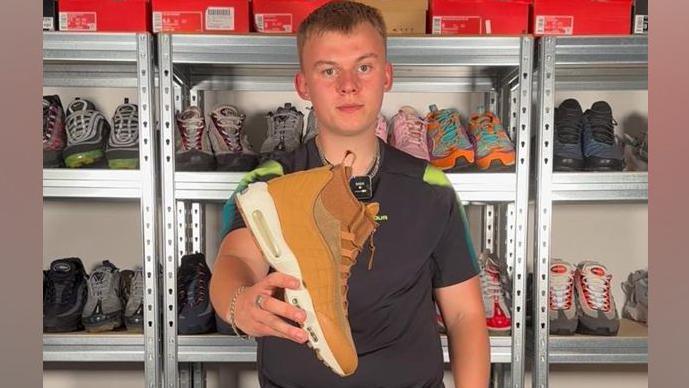 A young man with short dark blonde hair holding a mustard coloured boot out in front of him while standing in front of shelving units full of trainers