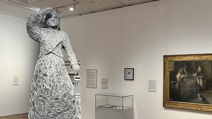 A wire-made sculpture of a woman in an art gallery, with paintings on the white walls behind it