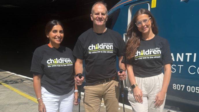 Two women and a man wearing black t-shirts with a white and green Railway Children writing and log on the front. They are standing on a station platform in front of a blue train. The woman on the left has black hair and white trousers and is smiling. The taller man has hiking poles in each hand and is wearing beige trousers. The woman on the right is younger, has long dark hair and transparent framed glasses. She is wearing beige trousers.