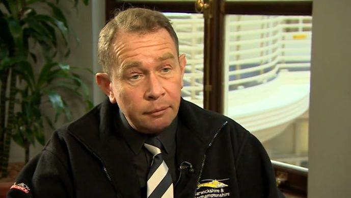 Philip Hollobone with short brown hair wearing a dark jacket with a yellow air ambulance logo, and a black and white striped tie.