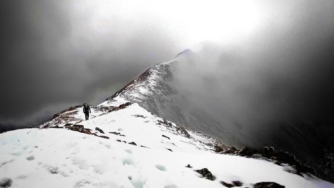 A man is tiny walking along the ridge of a snowy mountain. The picture is black and white.