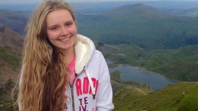 A teenage girl smiling at the camera, with long blonde hair, wearing a fur lined hoodie. She is standing high on the side of a mountain with other mountains, trees and a lake in the background.