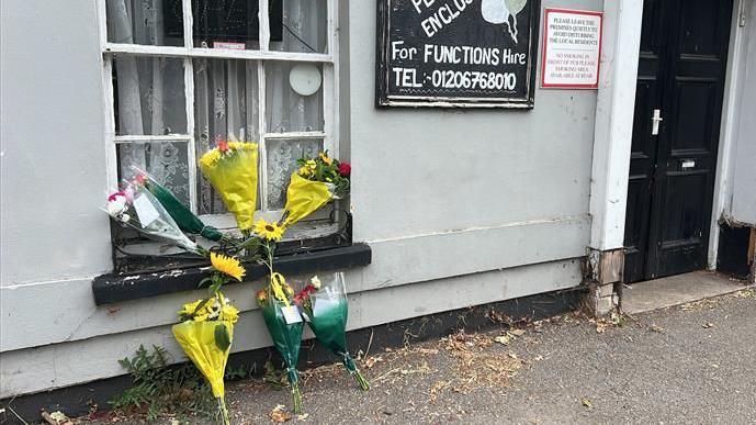 Flowers left outside the Cherry Tree pub. The pub is a grey building with a black door. The flowers have been left in front of a window.