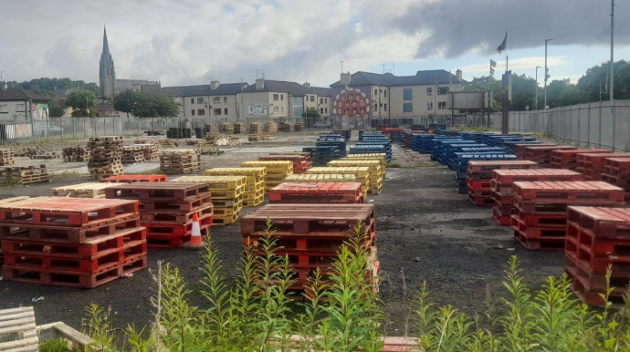Shows wooden pallets in red, yellow and blue with housing in the background and a cathedral with a large spire