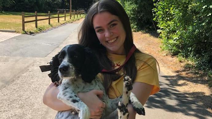 Maddy Lawrence. She has long dark hair and is wearing a yellow t-shirt. She is holding a black and white spaniel and is looking at the camera and smiling. Trees, a field and a pavement can be seen in the distance behind her.