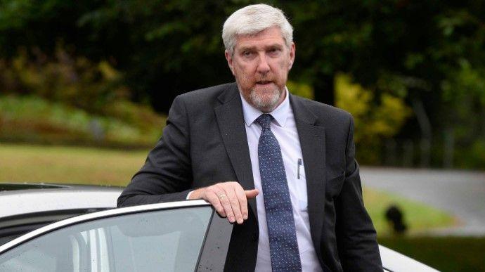 Shows John O'Dowd in a suit shirt and tie with his right hand resting on the passenger seat of a car