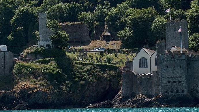 Dartmouth Castle seen from the sea with turrets and flags flying with a steep bank behind lined with trees.