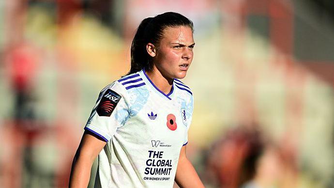 Close-up of Sarah Mayling during Aston Villa's Women's Super League match against Manchester United. She has dark hair, tied into a pony tail, and is wearing a white shirt which has purple trim on the collar and sleeves. There is a small Adidas logo on the right side and a main sponsor logo reading "The global summit on women & sport".