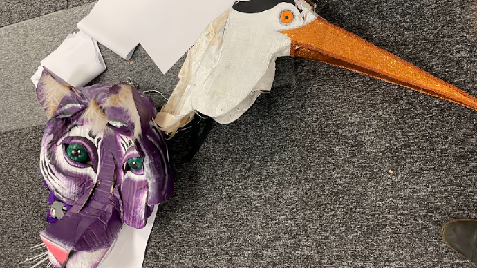 The head of a purple tiger puppet misshapen next to the damaged head of a crane-like animal lying on the floor.