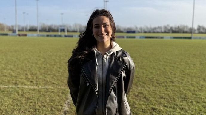 A woman with long dark hair and wearing a black leather jacket, standing on a rugby pitch and smiling at the camera. It's a sunny day and the sky is blue.