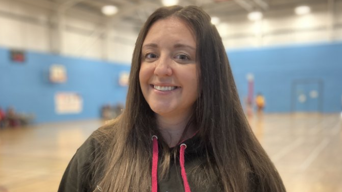 A woman with long dark hair smiles at the camera. She is wearing a hooded netball top, and is stood in a sports hall