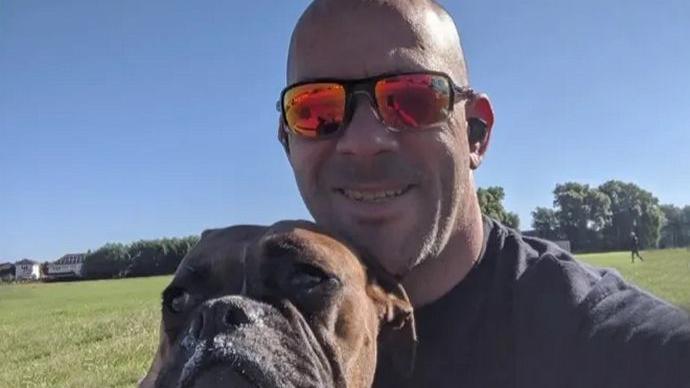 Glenn Cutter looking at the camera with his dog in front of him. The dog is a brown boxer-style breed and Mr Cutter has a shaved head and is wearing sunglasses