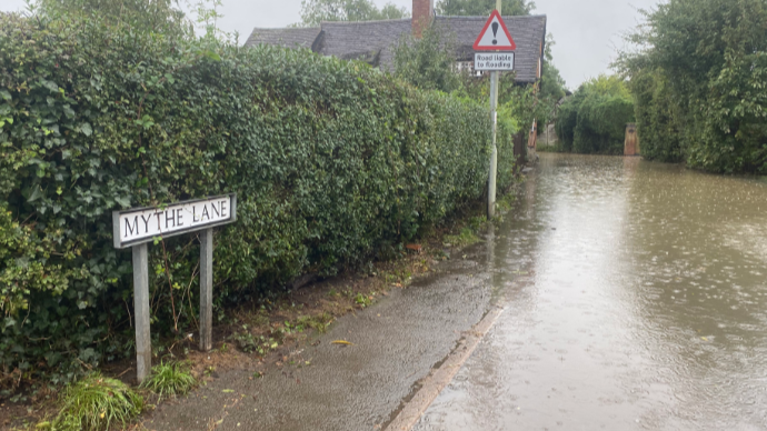 A hedgerow next to a road submerged with water and a house in the background