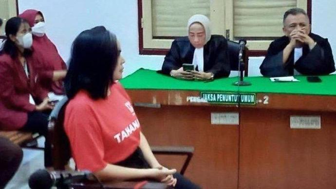 A woman in a red t-shirt sits in a chair in a courtroom in front of two judges, a man and a woman. The female judge looks at her mobile phone