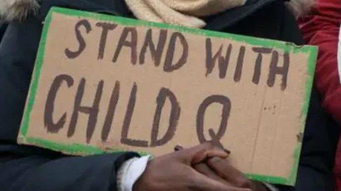 A person holds a cardboard sign which says Stand with Child Q