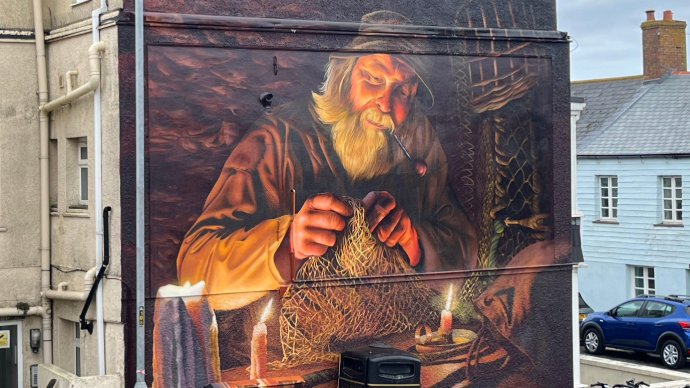 A mural on the side of a building depicts an older man with a long white beard, wearing a hat and smoking a pipe, as he mends a fishing net. He is illuminated by two candles in what appears to be a workshop or cabin