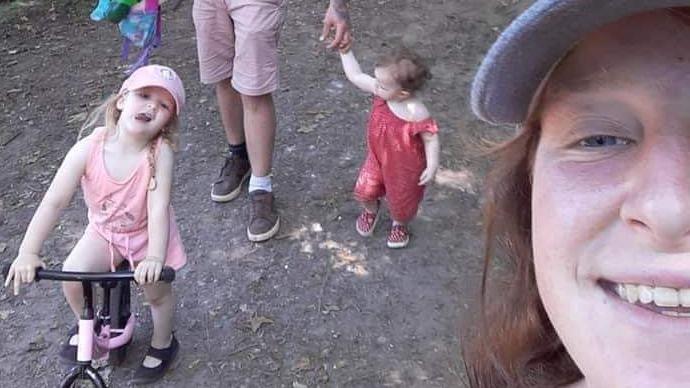 Alex Britton wearing a blue hat and smiling, and behind her are her two daughters, one wearing a pink dress and the other wearing a red outfit