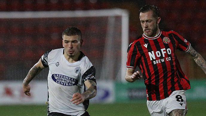Aidan Elliott-Wheeler (left) playing for Gateshead against Walsall in the FA Cup in pouring rain, with Walsall midfielder Charlie Lakin about to challenge him