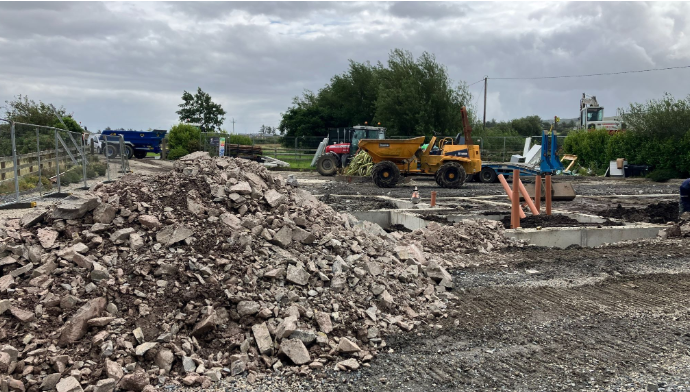 Shows a pile of bricks with a dumper truck and tractor in the background and some trees