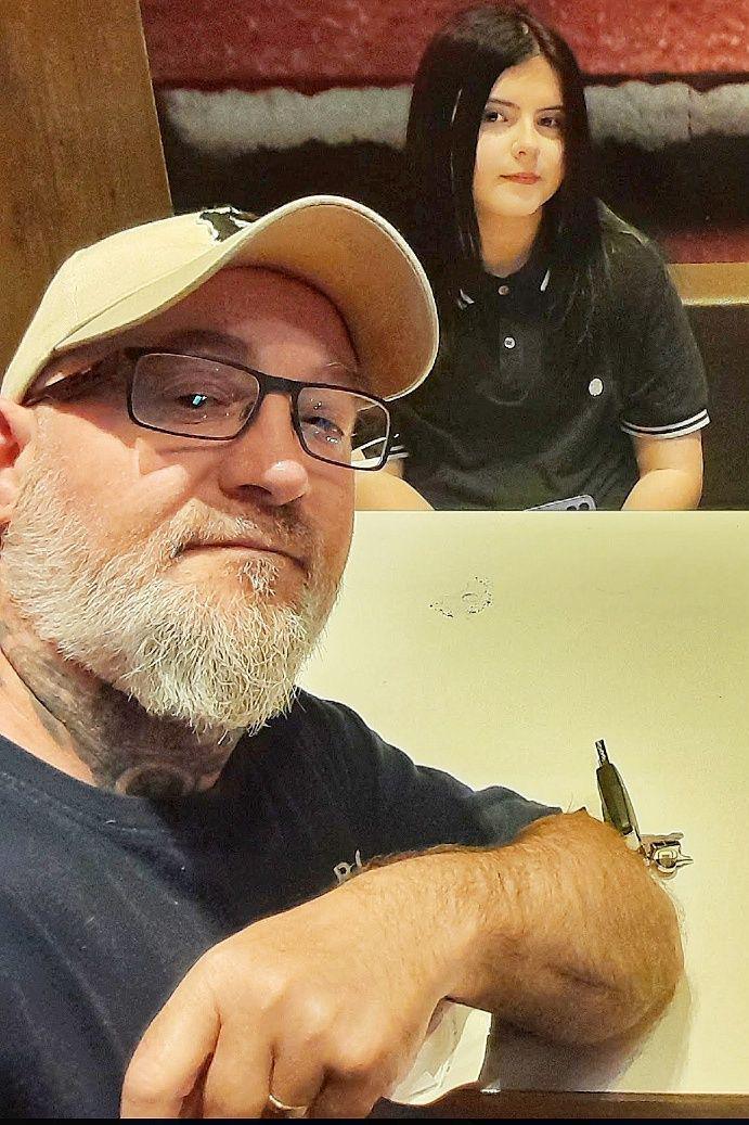 David Prosser is taking a selfie with his daughter in the background. They are sat at a table in what looks like a restaurant. He has a cream hat and glasses on, and is wearing a blue t-shirt. She has shoulder-length dark hair and has a black t-shirt on.