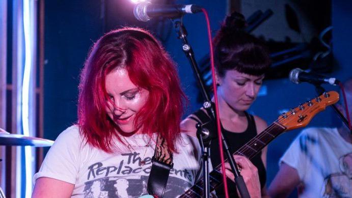 A woman with red hair plays guitar on stage wearing a white band T shirt for The Replacements. Behind her a woman in a black vest plays synth. She has black hair with a fringe.