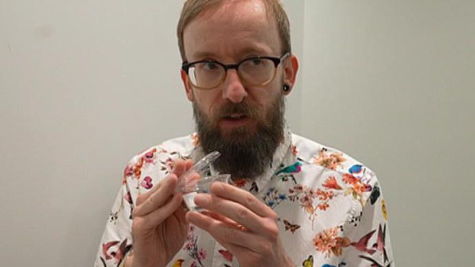 A man with glasses and a beard holding a plastic pot in his hands. He is wearing a watch and a shirt which has birds and flowers on.