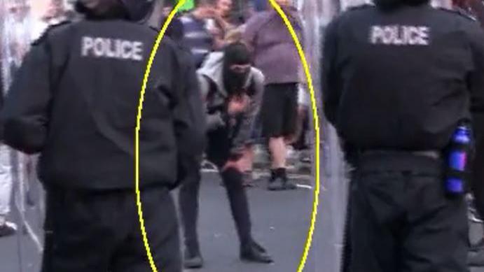 Still from body worn footage. Two police officers in riot gear have their backs to the camera. A man in black trainers and trousers bends over moving a black balaclava on his face