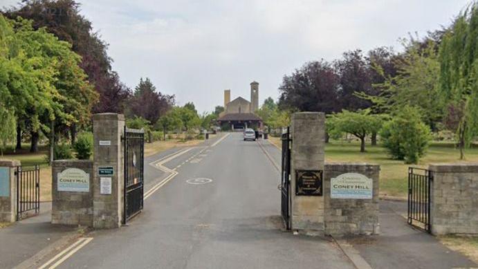 The entrance gates to Coney Hill Cemetery and Crematorium in Gloucester. Big stone posts with metal gates lead to a long driveway between green spaces with trees to the crematorium building.