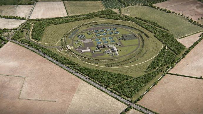 An aerial view of an artist's impression of a new waste treatment works in Cambridge. A circular area of land can be seen with grey buildings inside, surrounded by grass and trees. Beyond the perimeter are empty fields. 