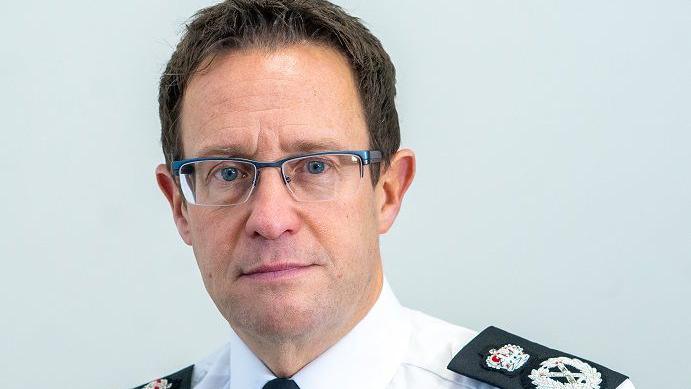 A man with short brown hair and glasses, wearing a white shirt with black police epaulets stands against a plain pale blue/grey background.