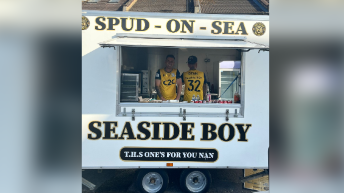 Max Linduschka and Bradley Gibbs are standing in a white food van which has Spud-on-Sea and Seaside Boy written on the front. They are both standing inside wearing yellow and black T-shirts.