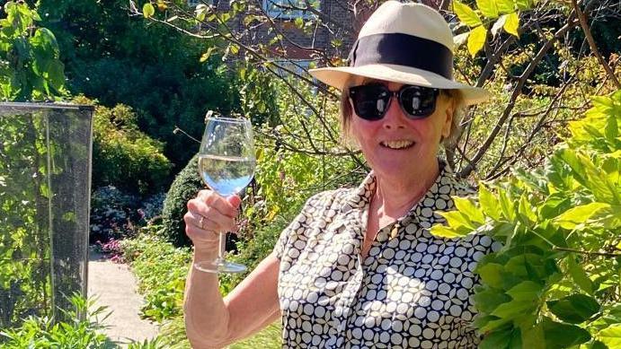 Sally Hall holds up a wineglass in a garden on a sunny day wearing black shades wearing a straw hat. 