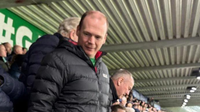 Minister Lyons standing in the stands at the match wearing a black puffer coat.