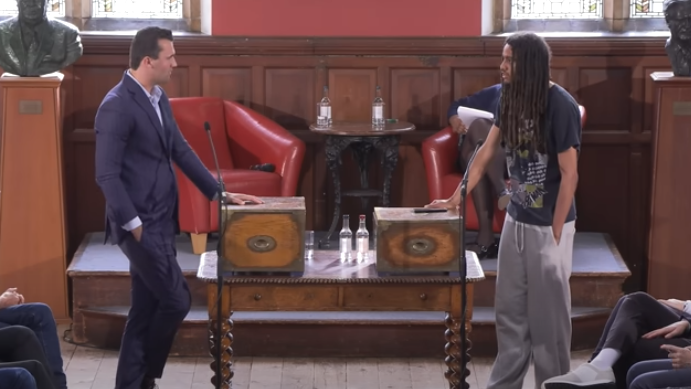 A still of Charlie Kirk and George Abaraonye debating at the Oxford Union. Kirk wears a dark blue suit. Abaraonye wears grey sweatpants and a black t-shirt. They are stood at an ornate wooden debating platform in a debating chamber.