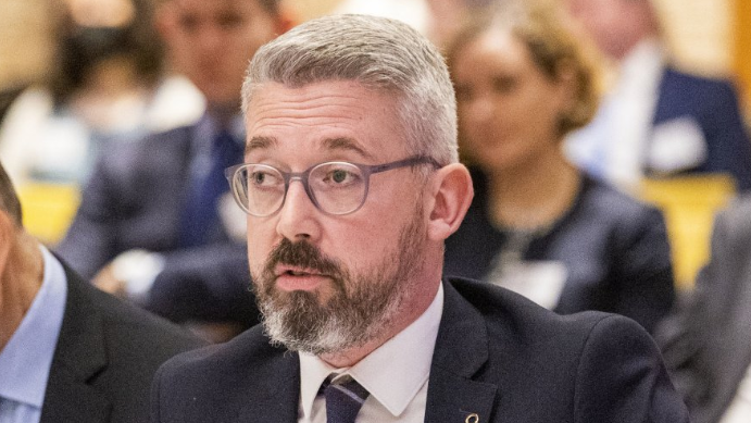 The former senator Niall Ó Donnghaile speaking at a committee meeting surrounded by other politicians. He is wearing a dark suit and tie with a white shirt.