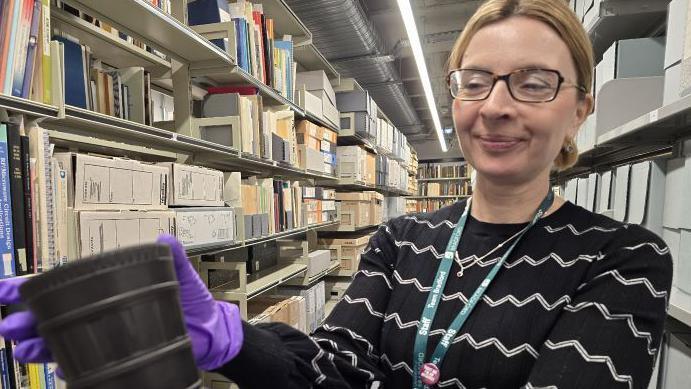 A woman with blonde hair pulled back into a ponytail wears dark-framed glasses and a black and white zig zag striped top. She is wearing a blue lanyard. She is wearing purple plastic gloves and holding a black vase and looking at it, smiling. She is stood between long rows of shelves, which have books, carboard folders and files on them.