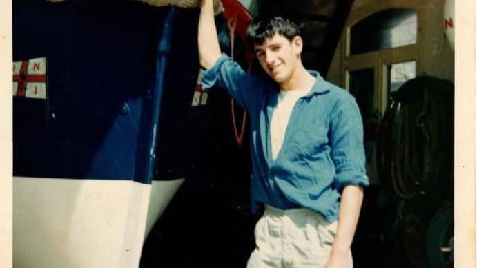 Richard when he was younger wearing a blue shirt and beige trousers leaning against an old RNLI boat smiling at the camera.
