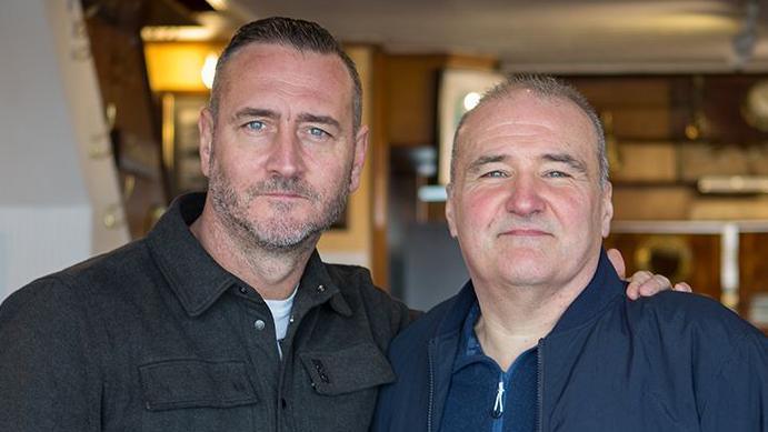 Actor Will Mellor, with his arm around the shoulder of Lee Castleton, the man he portrayed in Mr Bates vs the Post Office. Mellor has cropped dark hair and a short beard and is wearing a black jacket.