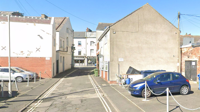 A narrow road between two sets of buildings with a car park on either side