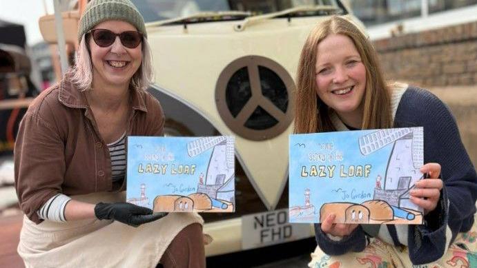 Jo Gordon and Hannah Graham holding a copy of The Story of the Lazy Loaf and smiling as they kneel in front of a cream-coloured van. The book cover is blue and shows bread loaves surrounded by Sunderland landmarks, such as Roker Pier and Fulwell Windmill. Jo has light pink-coloured hair, wears a green beanie, sunglasses, brown trousers and jacket. Hannah has long straight strawberry blonde hair and is wearing a blue cardigan.