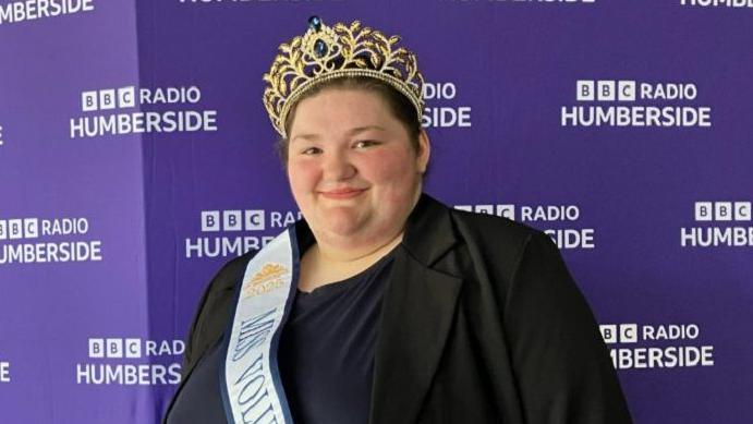 Philippa Mackinder wearing a sash and the gold crown she won as Miss Voluptuous England. She is wearing a black and navy top and is standing in front of the purple BBC Radio Humberside backdrop.